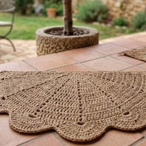 Può includere: Un tappeto semicircolare realizzato in fibra di juta naturale color marrone chiaro, caratterizzato da un dettagliato motivo all'uncinetto con bordo smerlato. Il tappeto presenta un design radiale che si irradia dal centro, con una trama tessuta.