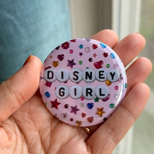 May include: A round pinback button featuring a light pink background decorated with various colorful, small, shiny star, heart, and flower-shaped embellishments. The text "DISNEY GIRL" is displayed in the center using white circular beads.