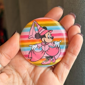 May include: A circular pinback button featuring a graphic of a cartoon mouse character wearing a pink dress, a matching pointed hat with a veil, and pink shoes. The background of the button consists of horizontal stripes in shades of orange, yellow, and blue.
