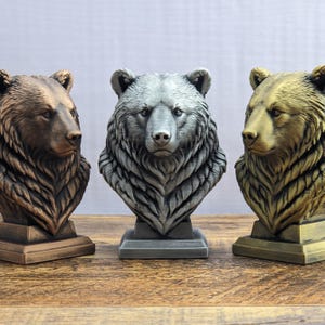 May include: Three bear head busts mounted on square bases. The left bust is finished in a metallic copper, the center bust is in a metallic silver, and the right bust is in a metallic gold. Each sculpture features detailed fur texture.