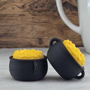 May include: Two small, matte black cauldron-shaped containers, each filled with a textured, bright yellow substance. The cauldrons feature small side handles and a rounded base, with one positioned upright and the other tilted slightly to the side.