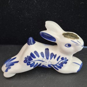 May include: A white ceramic rabbit figurine featuring blue hand-painted floral and leaf patterns. The rabbit is in a resting position with ears pointed upward. A gold-colored sticker on the ear reads MADE IN HOLLAND in black text.