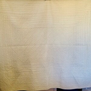 May include: A cream-colored, quilted blanket with a geometric pattern. The blanket features a grid-like design with horizontal and vertical stitching, creating a textured surface. The edges are neatly finished, and the overall aesthetic is simple and classic.