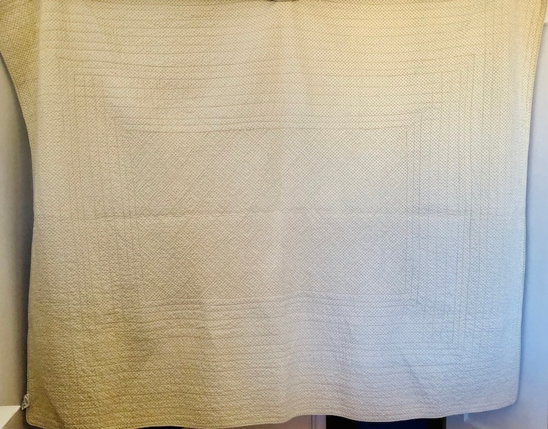 May include: A cream-colored, quilted blanket with a geometric pattern. The blanket features a grid-like design with horizontal and vertical stitching, creating a textured surface. The edges are neatly finished, and the overall aesthetic is simple and classic.