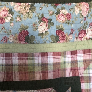 May include: A quilted textile featuring a floral pattern in shades of pink, white, and green on a light blue background. The quilt also has a plaid pattern in red, green, and white. The quilt is bordered with a solid green fabric.