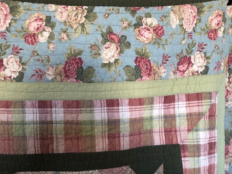 May include: A quilted textile featuring a floral pattern in shades of pink, white, and green on a light blue background. The quilt also has a plaid pattern in red, green, and white. The quilt is bordered with a solid green fabric.