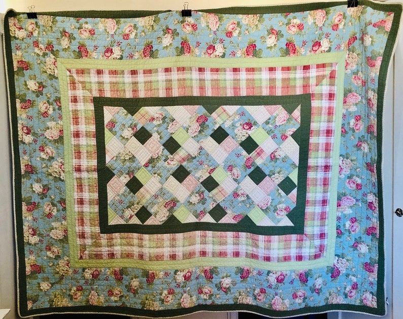 May include: A large, floral quilt with a light blue background and a repeating pattern of pink roses. The quilt features a green border and a central woven design with pink, green, and white squares. The overall design is a mix of floral and plaid patterns.