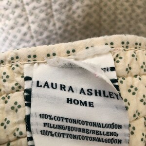 May include: Close-up of a cream-colored quilted fabric with a repeating pattern of small green dots. A white tag with the text "LAURA ASHLEY HOME" is attached to the fabric. The tag also includes the text "100% COTTON".