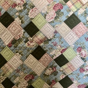 May include: A quilted textile featuring a patchwork design. The pattern incorporates squares of various fabrics, including floral prints with pink and white roses, light blue backgrounds, and solid dark green and light green squares. The quilt has a diamond pattern.