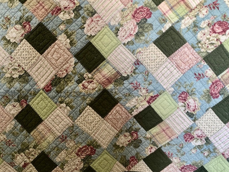 May include: A quilted textile featuring a patchwork design. The pattern incorporates squares of various fabrics, including floral prints with pink and white roses, light blue backgrounds, and solid dark green and light green squares. The quilt has a diamond pattern.