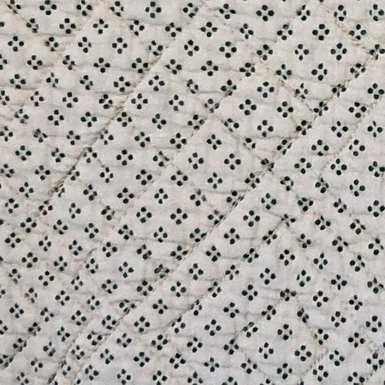 May include: Close-up of a white fabric with a repeating pattern of small, black floral-like shapes. The fabric is quilted with diagonal stitching, creating a diamond pattern across the surface.