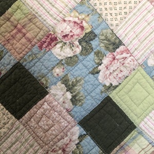 May include: Close-up of a patchwork quilt with a floral pattern. The quilt features squares of various colors and patterns, including light blue with pink and white flowers, green, and striped fabric. The quilt is quilted with a diamond pattern.