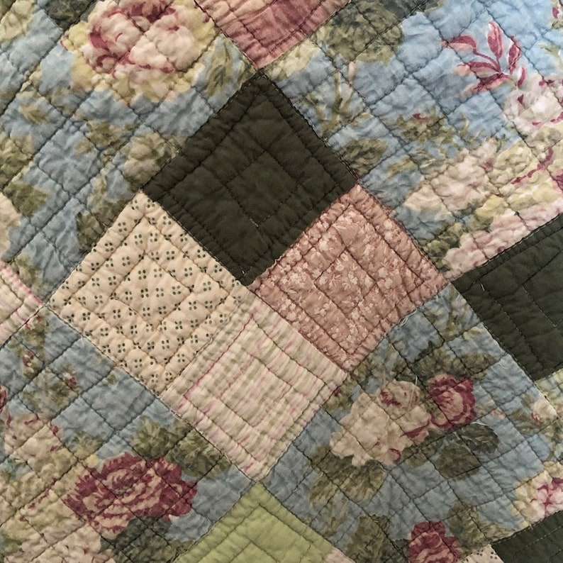 May include: Close-up of a quilted fabric with a patchwork design. The quilt features squares in various colors, including light blue, green, and pink, with floral patterns and geometric shapes. The stitching creates a textured surface.