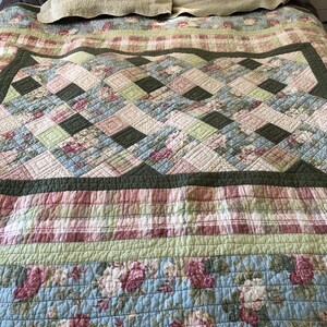 May include: A quilted bedspread with a floral and geometric pattern. The quilt features a combination of light blue, pink, green, and dark green squares and stripes. The top of the bedspread has a pillow.