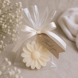 Puede incluir: Un artículo de cera en forma de margarita de color crema empaquetado en una bolsa de plástico transparente. La bolsa está atada con una cinta de satén color crema y tiene una etiqueta de papel marrón con el texto Thank you en negro.