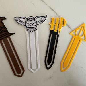 Peut inclure: Quatre marque-pages plats en plastique. Le premier est marron en forme de chapeau pointu. Le deuxième est blanc avec un hibou tenant une enveloppe. Le troisième est noir avec des lettres HP jaunes. Le quatrième est jaune en forme de triangle.