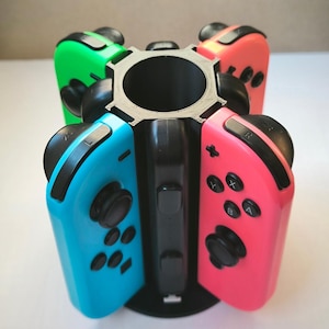 Può includere: Un supporto di ricarica in plastica nera progettato per contenere quattro controller di gioco. Il supporto presenta una colonna cilindrica centrale con fessure per agganciare controller in verde neon, rosa neon, blu neon e rosso neon, con tasti neri.