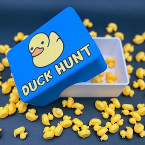 May include: A blue rectangular box with a yellow duck graphic and the text DUCK HUNT printed on the lid, partially open to reveal a white interior, surrounded by numerous small yellow rubber duck figurines scattered across the surface.