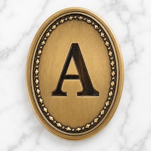 May include: An oval-shaped brass plaque featuring a prominent capital letter A in the center. The plaque has a brushed metallic finish and is bordered by a decorative rim consisting of small, repeating diamond-shaped studs and circular beads.