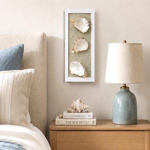 May include: A vertical rectangular white frame containing three white oyster shells with gold-painted edges, mounted on a textured tan burlap background. The shells are arranged in a single column, showcasing their natural interior pearlescent finish.