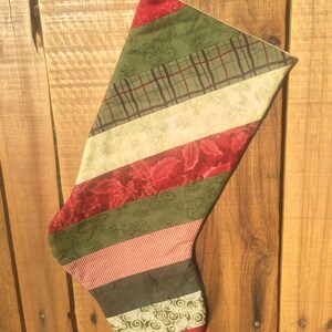 May include: A Christmas stocking constructed from diagonal fabric strips in various patterns and colors, including red holly prints, olive green floral motifs, dark green solids, cream swirls, and thin red and white stripes, with a hanging loop.