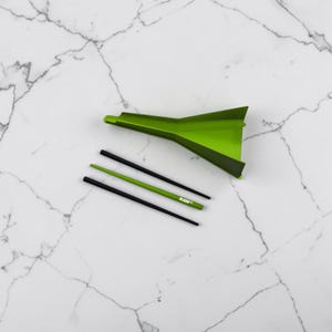 May include: A lime green plastic funnel positioned above three thin sticks. Two sticks are black and one is lime green with the text RAW printed in white. The funnel has a wide opening at one end and a narrow spout at the other, designed for transferring small materials.
