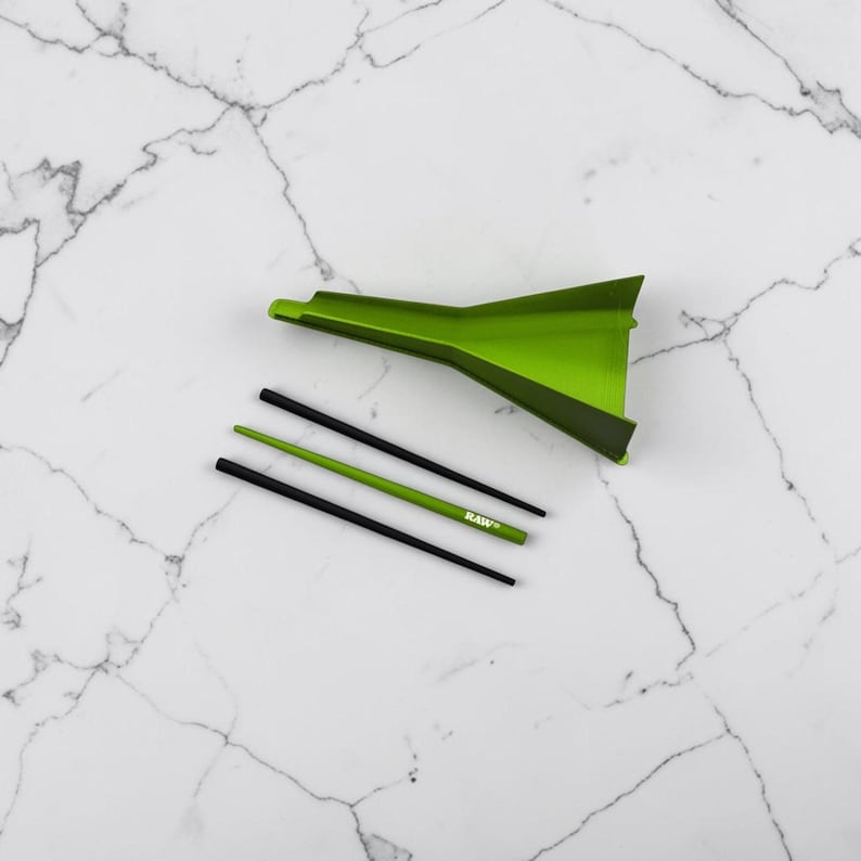 May include: A lime green plastic funnel positioned above three thin sticks. Two sticks are black and one is lime green with the text RAW printed in white. The funnel has a wide opening at one end and a narrow spout at the other, designed for transferring small materials.