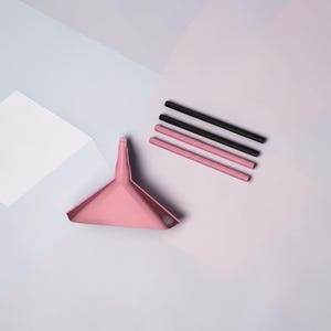 May include: A pink triangular funnel with a narrow spout positioned next to two black cylindrical straws and two pink cylindrical straws arranged in a parallel row. The items are displayed on a flat surface with geometric shapes in shades of pink and blue.