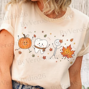 Puede incluir: Una camiseta de manga corta de color crema con un estampado gráfico de una calabaza sonriente, un diente sonriente con bufanda, un espejo dental y una hoja de otoño, todo rodeado de pequeñas hojas que caen y estrellas.
