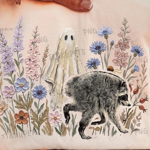 Puede incluir: Una camiseta de color claro con una ilustración impresa de un mapache gris caminando junto a una figura de fantasma blanca, rodeado de flores silvestres coloridas en tonos morado, rosa, azul y naranja, con una marca de agua repetida.