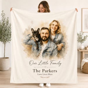 May include: A cream-coloured throw blanket featuring a watercolour-style portrait of a man, a woman, and a French Bulldog. The text reads: Our Little Family, The Parkers, Love Lives Here. The design is centred on the fabric with soft, blended edges.