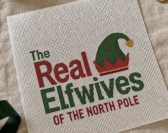 Może przedstawiać: Kwadratowe płótno do haftu z napisem The Real Elfwives OF THE NORTH POLE wykonanym czerwoną i zieloną nicią. Zielono-czerwona czapka elfa z żółtym pomponem jest wyhaftowana nad słowem Elfwives na białej tkaninie o splocie siatkowym.