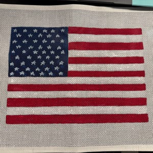 May include: A rectangular needlepoint canvas featuring a printed design of the United States flag. The design includes a blue canton with white star outlines and alternating horizontal red and white stripes on a white grid mesh background.