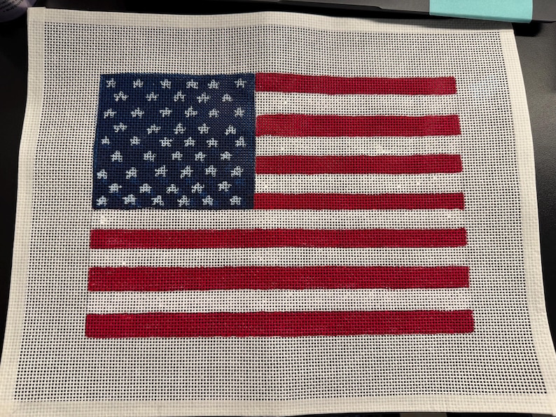 May include: A rectangular needlepoint canvas featuring a printed design of the United States flag. The design includes a blue canton with white star outlines and alternating horizontal red and white stripes on a white grid mesh background.