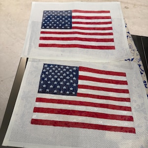 May include: Two rectangular needlepoint canvases featuring the American flag design. Each canvas displays a blue field with white stars in the upper left corner and alternating horizontal red and white stripes across the remaining surface area.