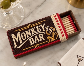 Może przedstawiać: Prostokątne drewniane pudełko na zapałki z haftowaną nakładką na ciemnobrązowym tle, kremowym napisem MONKEY BAR oraz ilustracją żółtej małpy trzymającej kieliszek do martini. Pudełko zawiera drewniane zapałki z czerwonymi końcówkami.