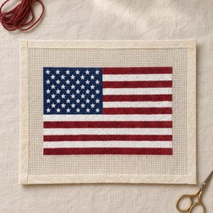 May include: A rectangular needlepoint canvas featuring a cross-stitched American flag design. The flag displays a blue field with white stars and alternating red and white horizontal stripes, centered on a plain, light-colored grid fabric.