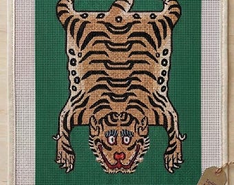 Może przedstawiać: Płótno do haftu przedstawiające beżowego tygrysa w czarne paski na jednolitym zielonym tle. Tekst EMERALD TIBETAN TIGER - 13 MESH - CANVAS ONLY jest nadrukowany u góry, a HAND-PAINTED IN NYC u dołu. Dołączona jest mała brązowa etykieta.