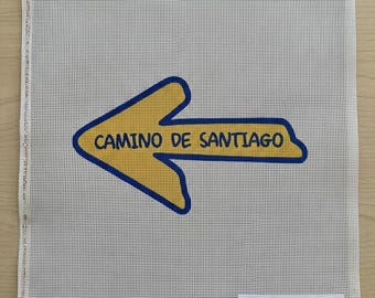 May include: A square needlepoint canvas featuring a yellow arrow pointing to the left with a blue outline. The text CAMINO DE SANTIAGO is printed in black inside the arrow. A small white label in the bottom right corner reads SANTIAGO ART CANVASES - CAMINO ARROW #001.