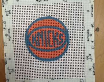 Może przedstawiać: Kwadratowe płótno do haftu z centralną grafiką pomarańczowej piłki do koszykówki i niebieskim napisem KNICKS wyhaftowanym pośrodku. Płótno jest wykończone białą lamówką z nadrukiem małych czarnych ikon kotów oraz napisem CAT LOVER.