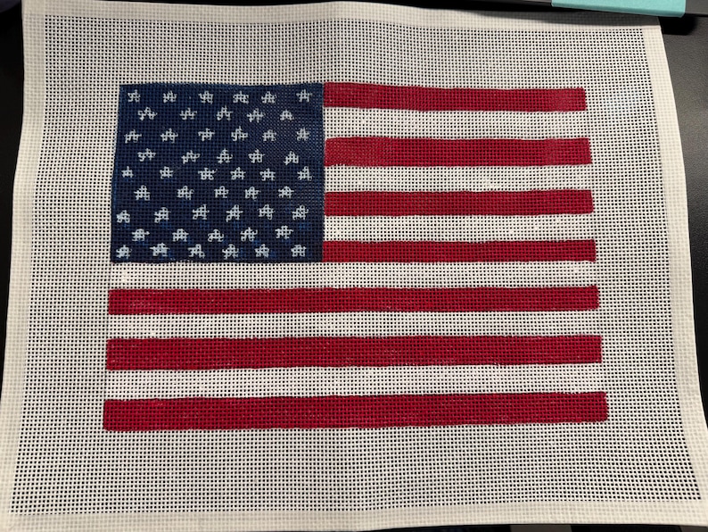 May include: A needlepoint canvas featuring a printed design of the United States flag. The design includes a blue canton with white star outlines and alternating horizontal red and white stripes, all set against a white grid mesh background.