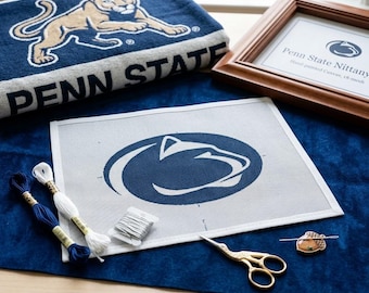 Może przedstawiać: Płótno do haftu z logo lwa Nittany z Penn State, w towarzystwie dwóch motków nici do haftowania, szpulki nici, złotych nożyczek i magnesu na igły. W pobliżu znajduje się oprawiony wydruk oraz granatowy ręcznik z tym samym logo.