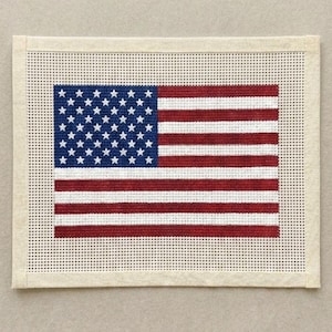 May include: A rectangular needlepoint canvas featuring the United States flag design. The flag consists of a blue canton with white stars and alternating red and white horizontal stripes, all set against a plain, unstitched cream-colored border.