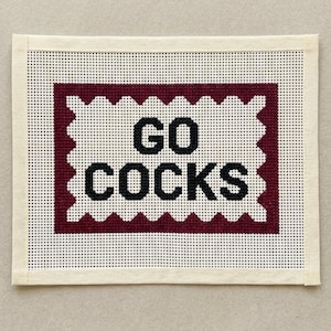 May include: A rectangular needlepoint canvas featuring a white center with the black text GO COCKS, surrounded by a dark maroon scalloped border. The design is set against a white grid background with finished cream-colored fabric edges on all sides.