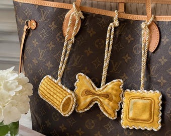 May include: A brown monogram tote bag featuring three hanging ornaments shaped like pasta pieces. The ornaments include a yellow rigatoni, a farfalle bow tie, and a square ravioli, each suspended by a twisted white and gold rope cord.