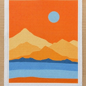 May include: A rectangular needlepoint canvas featuring a landscape design with a bright orange sky, a light blue sun, layered yellow and orange mountains, a dark blue mountain range, a light blue lake, and an orange foreground. Text reads TAYLOR & KOBI TK-001.