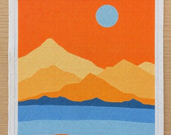 May include: A needlepoint canvas featuring a landscape design with a bright orange sky, a light blue sun, layered yellow and orange mountains, a dark blue mountain range, and a blue water foreground. The text TAYLOR & KOBI and TK-001 are printed.