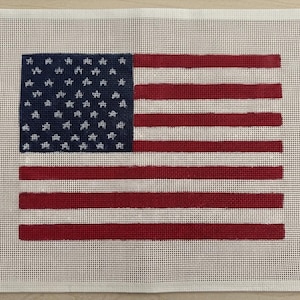May include: A rectangular needlepoint canvas featuring a printed design of the American flag. The flag consists of a dark blue canton with white star outlines and alternating horizontal red and white stripes, all set against a white mesh background.