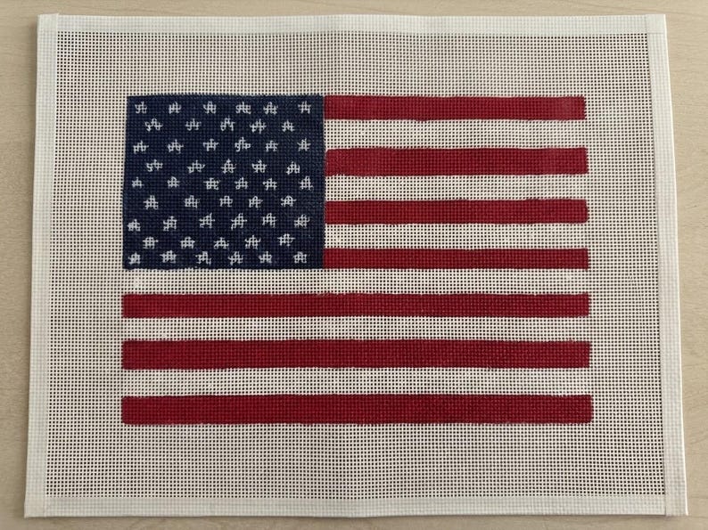 May include: A rectangular needlepoint canvas featuring a printed design of the American flag. The flag consists of a dark blue canton with white star outlines and alternating horizontal red and white stripes, all set against a white mesh background.