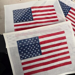 May include: Two rectangular needlepoint canvases featuring a printed American flag design. Each canvas displays a blue field with white stars and alternating red and white horizontal stripes on a white mesh grid background, ready for embroidery.